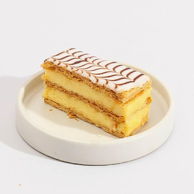 Kue lapis krim kuning dengan hiasan cokelat, disajikan di piring putih, online florist